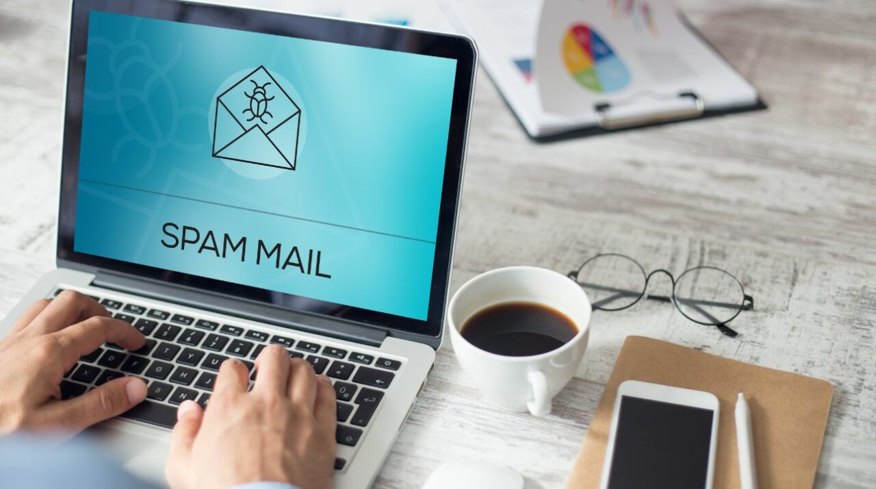 two hands on laptop keyboard looking for the origin of the word spam with spam mail written on screen on blue background and cup of coffee and phone nearby