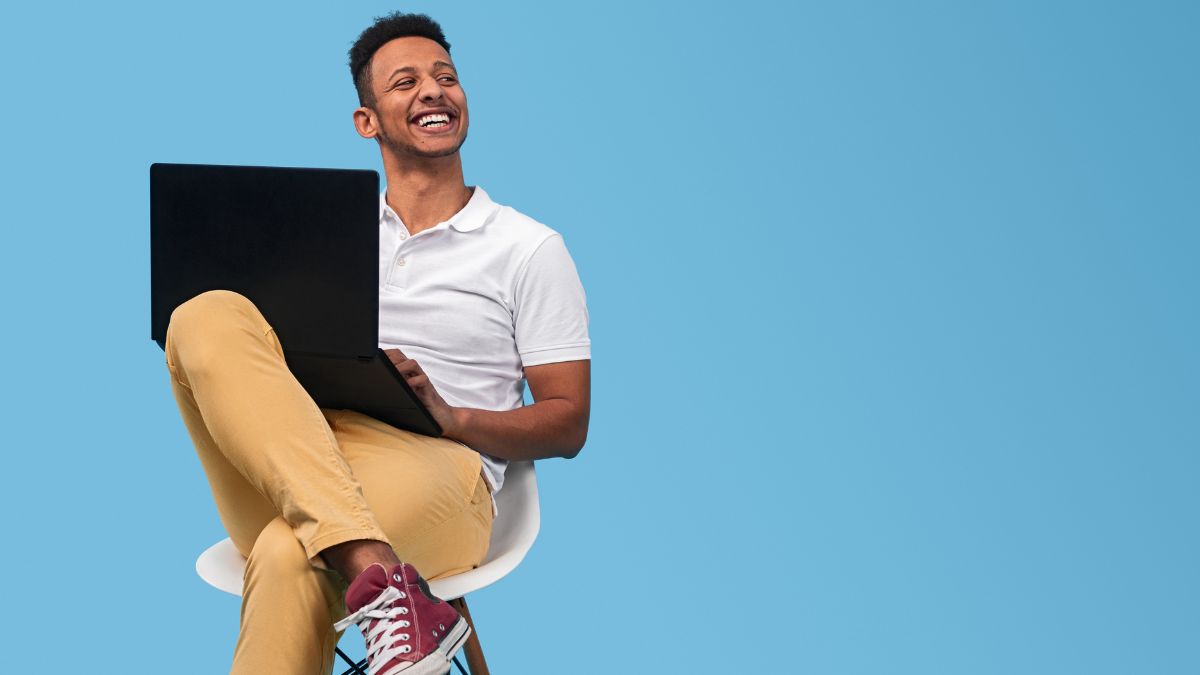 Young man in khaki pants and a short sleeve shirt looks at his laptop on a light blue background.
