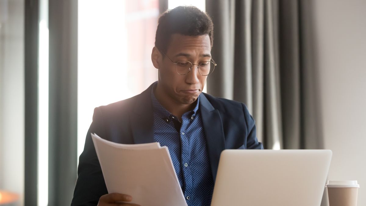 Young, bespectacled man looks at laptop and papers while dealing with email fatigue.