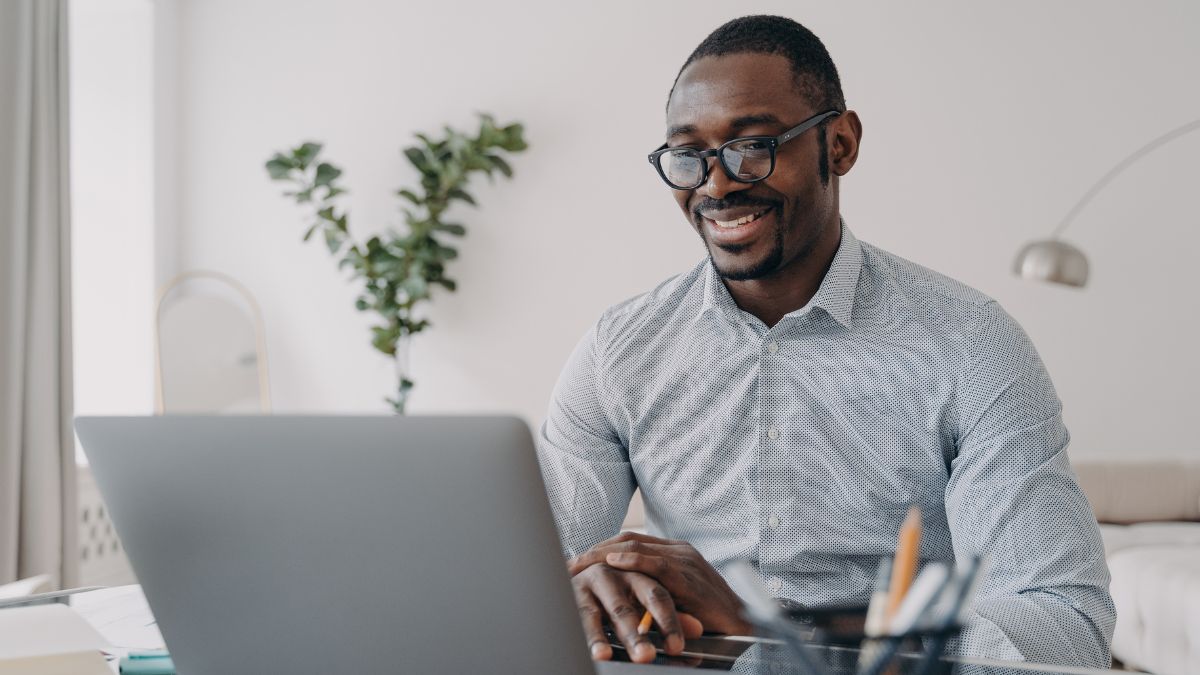Young spectacled man sits at laptop to do some cold emailing while wearing a long sleeve dress shirt.